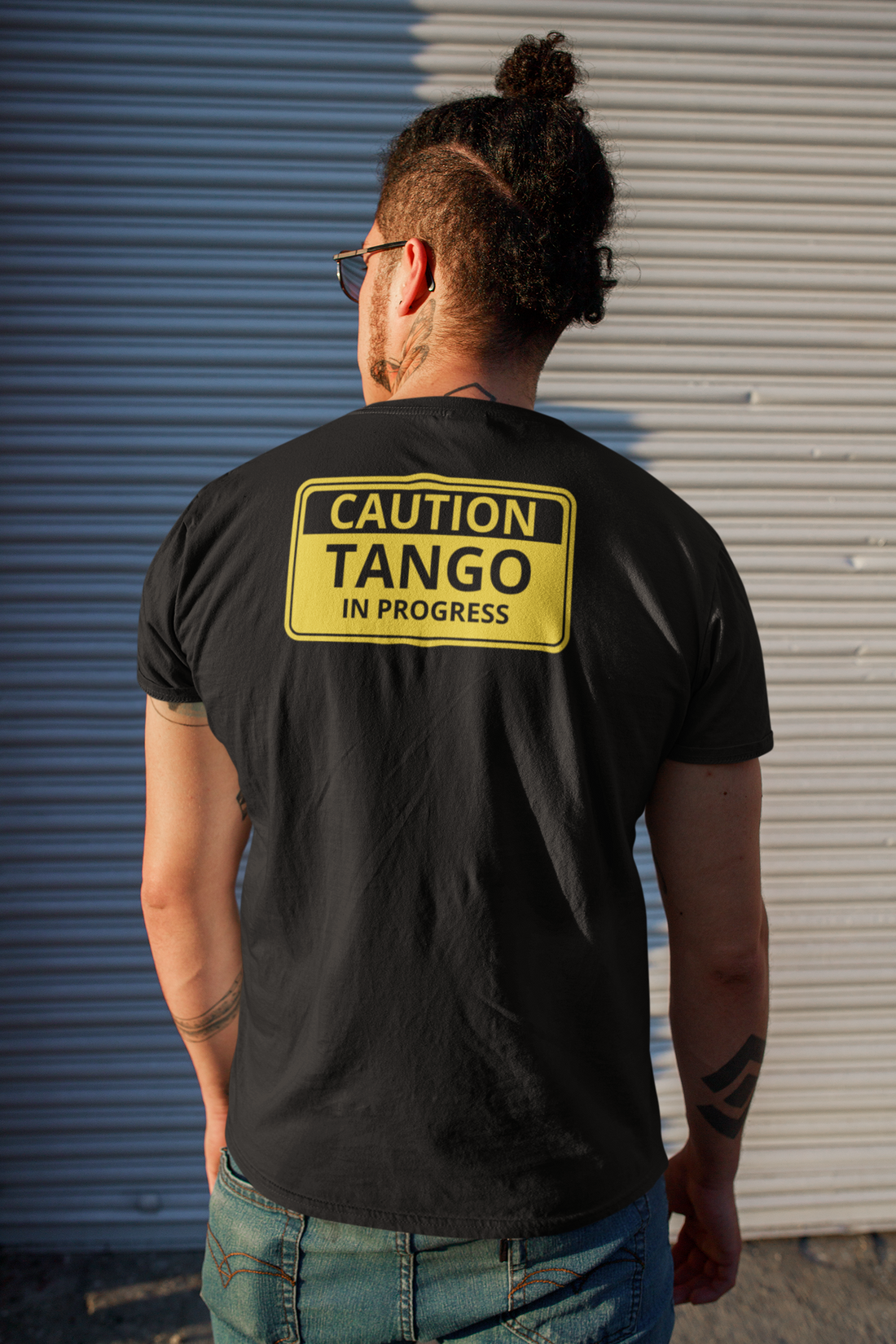 A male wearing a black T-shirt with the text "Caution, Tango in Progress" on top of a yellow and black warning sign