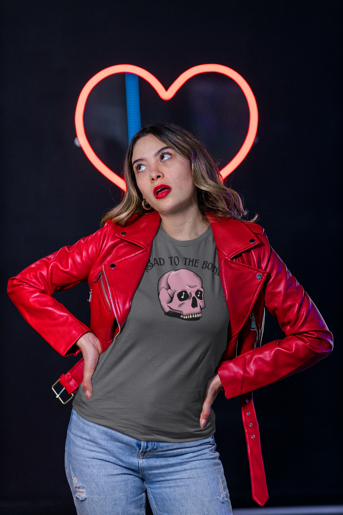 A female standing in fornt of a neon heart, wearing a grey music tshirt with the text "bad to the bones" and the image of a pink skull