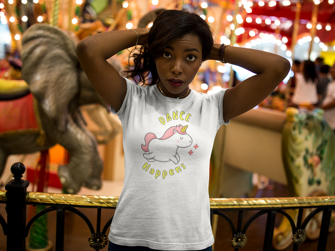 A female wearing a white Tshirt design with a happy and dancing unicorn with the text "dance happens"
